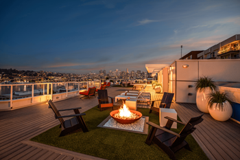 A rooftop patio with a fire pit and chairs overlooking a city skyline at dusk.